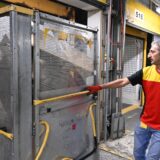 عامل يرتدي زي شركة (DHL) اللوجستية يقوم بسحب حاوية طرود معدنية كبيرة داخل مستودع أو مركز شحن.