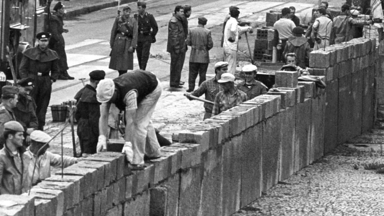 صورة من أرشيف - ١٨ أغسطس/آب ١٩٦١، ألمانيا الشرقية في برلين: تحت إشراف ضباط شرطة الشعب المسلحين، يقوم فريق من عمال البناء في برلين الشرقية بتشييد جدار على حدود القطاع السوفيتي الأمريكي في ساحة بوتسدام. 
Foto: UPI/ dpa

