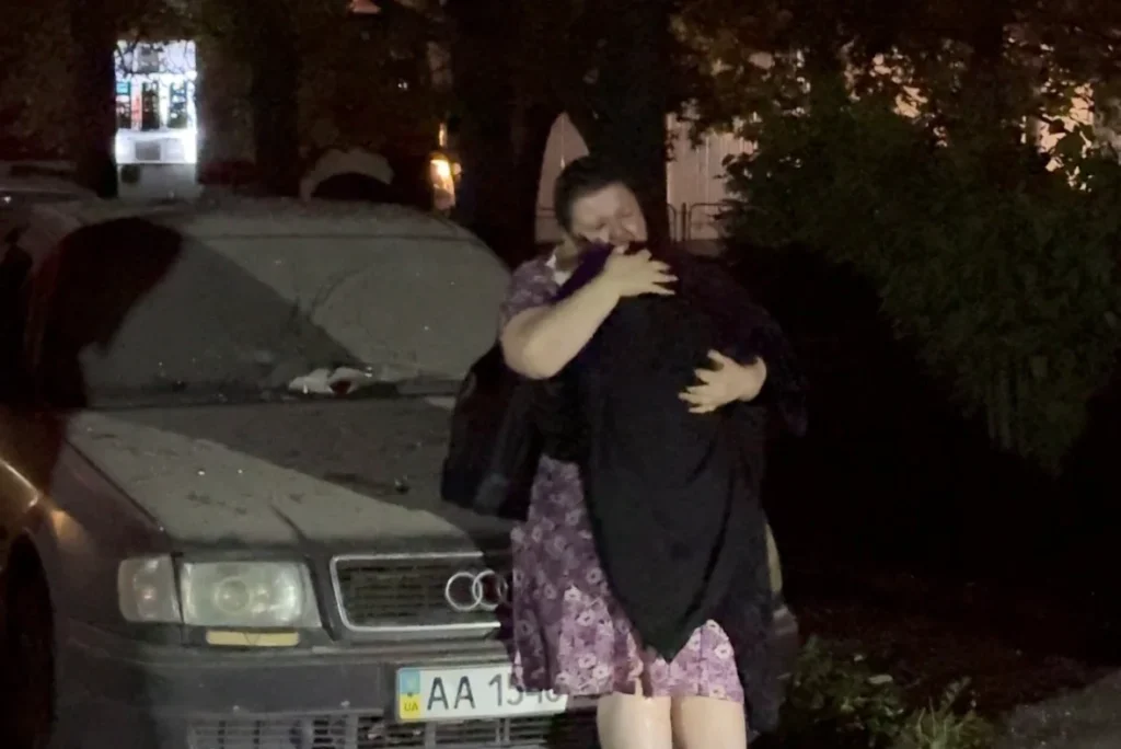 Eine Frau lehnt an eine Motorhaube und hält ein in eine Decke gewickeltes Tier.