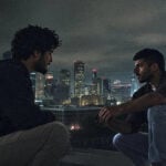 Das Bild ist ein Still aus dem Film Naza. Es zeigt zwei Männer im Dunkeln. Im Hintergrund sind Hochäuser zu sehen