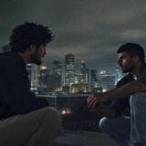 Das Bild ist ein Still aus dem Film Naza. Es zeigt zwei Männer im Dunkeln. Im Hintergrund sind Hochäuser zu sehen
